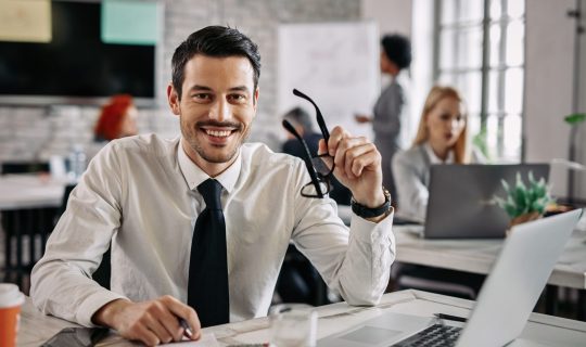 Young happy businessman working in the office and looking at camera. There are people in the background.