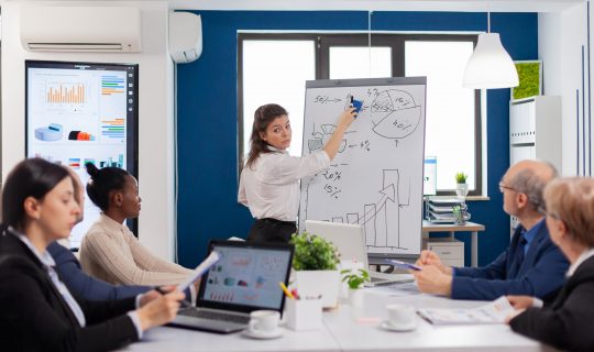 Company head standing in front of audience diverse staff members making presentation. Serious speaker boss executive, business trainer explaining development strategy to motivated mixed race employees.