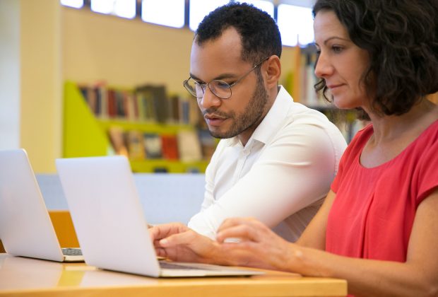 Two concentrated students talking and looking at laptop at library. Focused people studying with laptops together. Technology, education concept