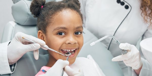 Happy afro american girl sitting in stomatologist chair with open mouth while professional dentist doing regular check up of teeth using dental probe and mirror. Female nurse assisting.